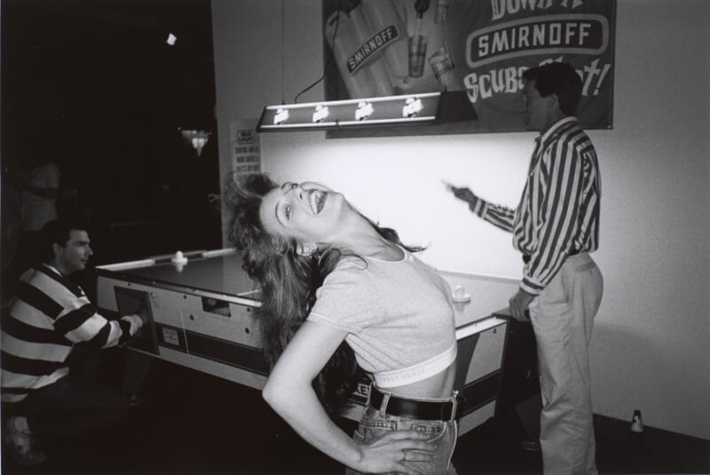 A woman laughs with her head tilted back in front of an air hockey table, while two men stand nearby in a dimly lit room with a Smirnoff sign on the wall.
