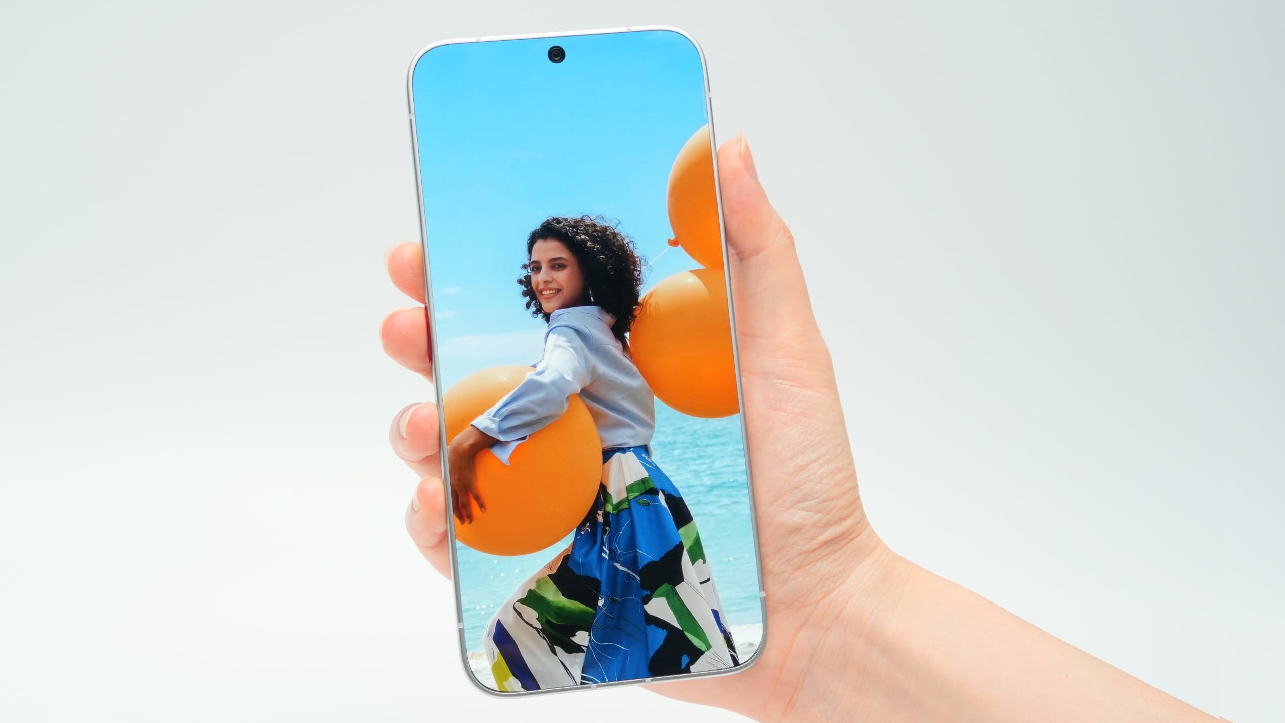 A hand holds a smartphone displaying a portrait of a woman with curly hair holding large orange balloons against a blue sky.