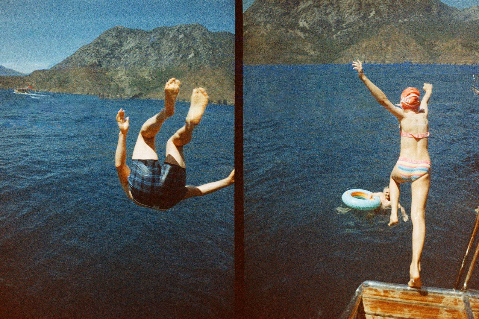 Two people jump off a dock into a blue lake; one is mid-flip in swim trunks, the other leaps toward a float, wearing a striped swimsuit, with mountains in the background.