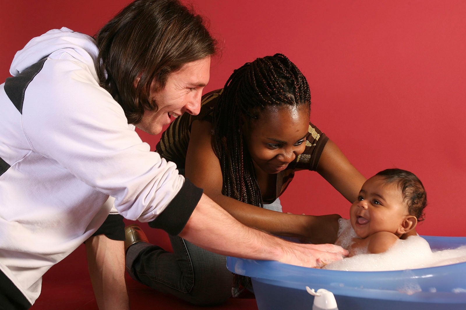 Two adults smile as they bathe a happy baby in a blue tub filled with bubbles against a red background.