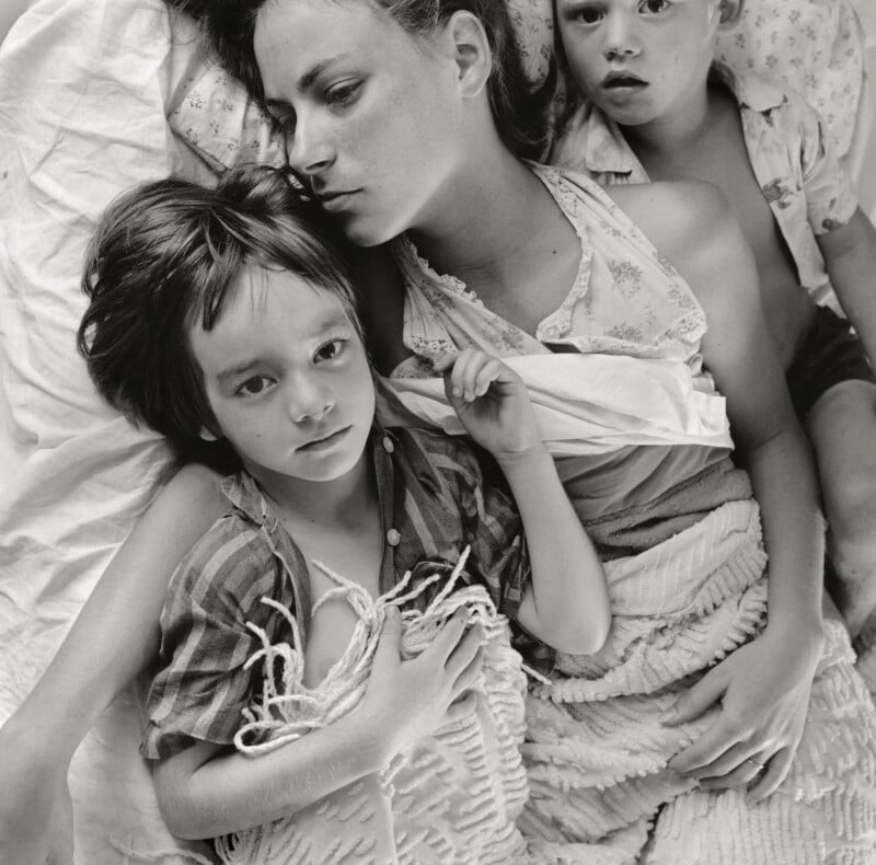 A woman lies in bed with her arms around two children, all looking thoughtful or somber. They are partially covered by blankets, close together, and sharing an intimate, quiet moment.