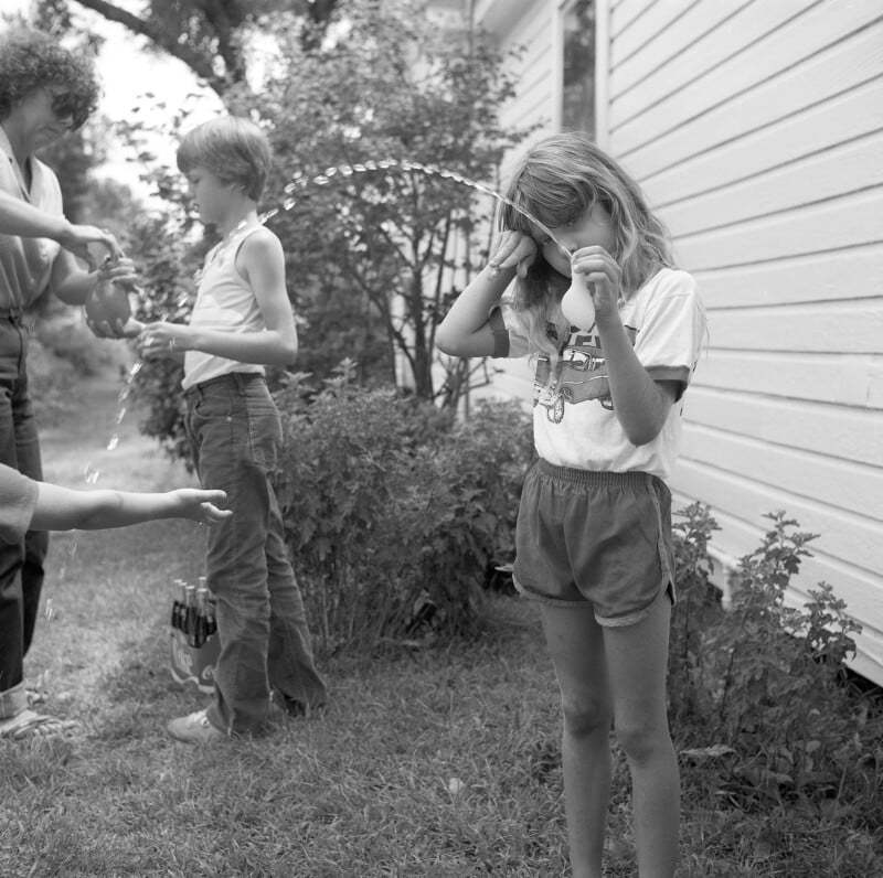 A girl in shorts shields her face while holding a small water balloon. Two children and an adult prepare more balloons nearby. Water splashes in the air beside a white house.