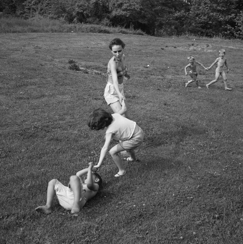 A woman pulls a child up from the grass while another child helps. Two more children run while holding hands in the background on a grassy field.
