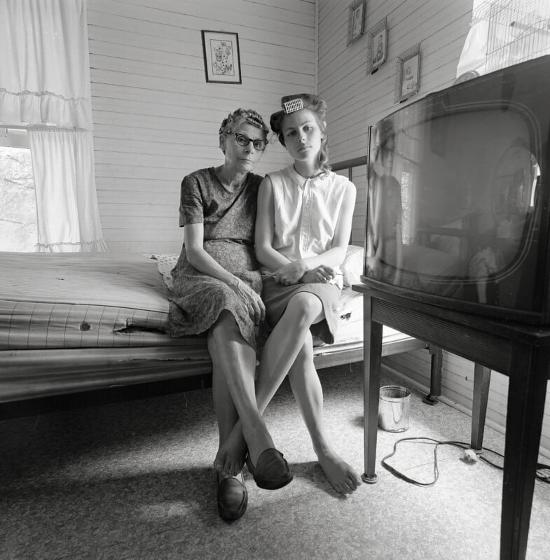 Two women sit close together on a bed in a modest room, looking seriously at the camera. One wears glasses; the other has hair rollers. A large old TV is in the foreground, and framed pictures hang on the white-paneled wall.
