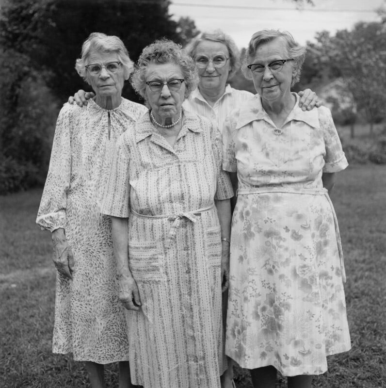 Four elderly women stand close together outdoors, wearing patterned dresses and glasses. They pose for a black-and-white photo, with trees and grass visible in the background.