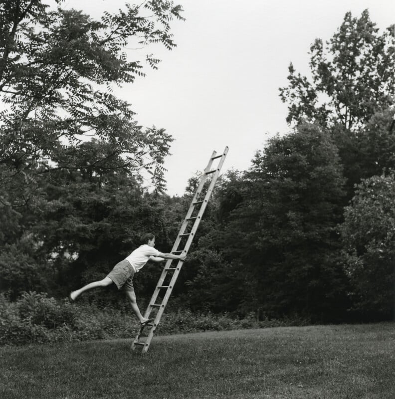 A person in shorts and a t-shirt balances on one leg while leaning against a tall ladder in a grassy area surrounded by trees.