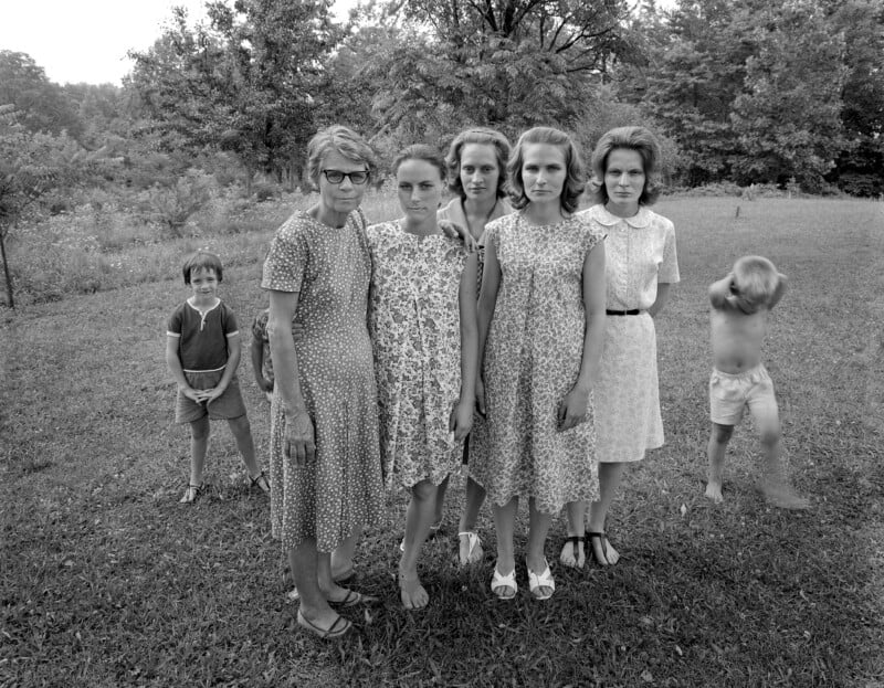 Six women in dresses stand in a grassy yard, facing the camera. Two children are in the background, one running and blurry, the other standing still. Trees and greenery fill the background.