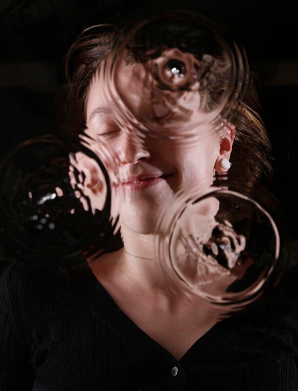 A woman stands behind rippled glass, her face partially distorted by three circular patterns, eyes closed and smiling softly, wearing a black top and pearl earrings against a dark background.