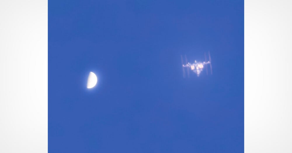 A blurred photo shows the International Space Station passing near a half moon in a blue sky during daylight.