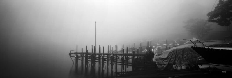 A wooden pier extends into a calm, foggy lake, with a covered boat docked in the foreground.