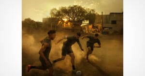 Three men play soccer on a dusty field at sunset, kicking up dirt as they chase the ball. The warm light and silhouettes of trees and buildings create a dramatic, action-filled scene.