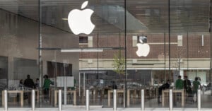 Apple Store with large glass windows, white Apple logos, and several employees in green shirts inside. Wooden tables are arranged in rows, and the outside shows reflections of buildings and trees.
