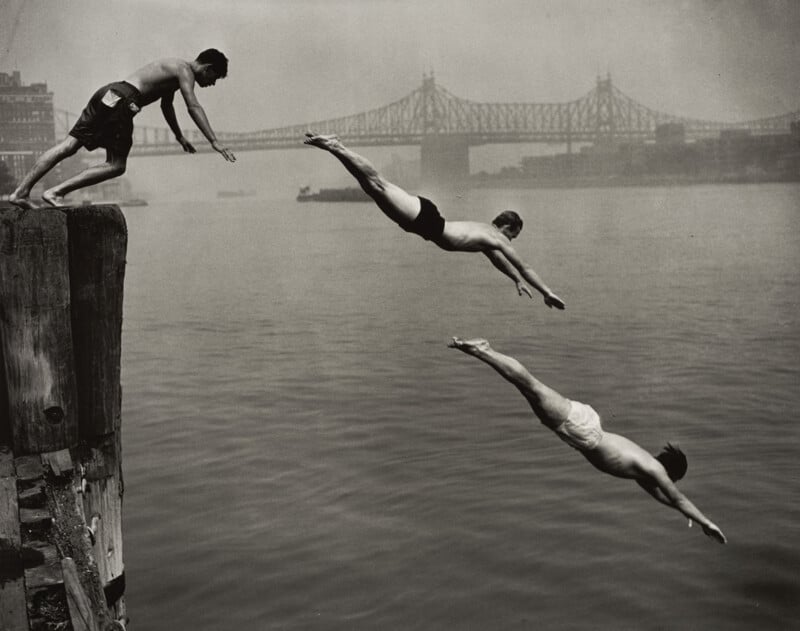 Three men dive into a river from a wooden pier, with a large bridge visible in the background.