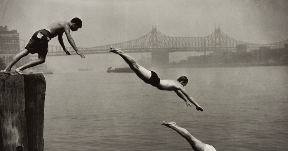 Three men dive into a river from a wooden pier with a large bridge visible in the background.