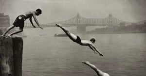 Three men dive into a river from a wooden pier with a large bridge visible in the background.