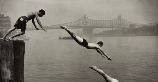 Three men dive into a river from a wooden pier with a large bridge visible in the background.