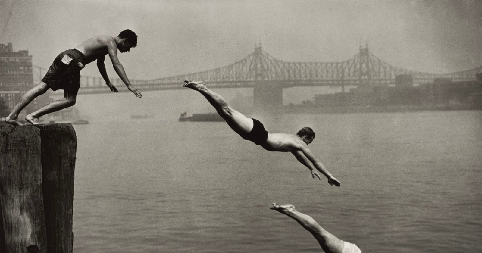 Three men dive into a river from a wooden pier with a large bridge visible in the background.