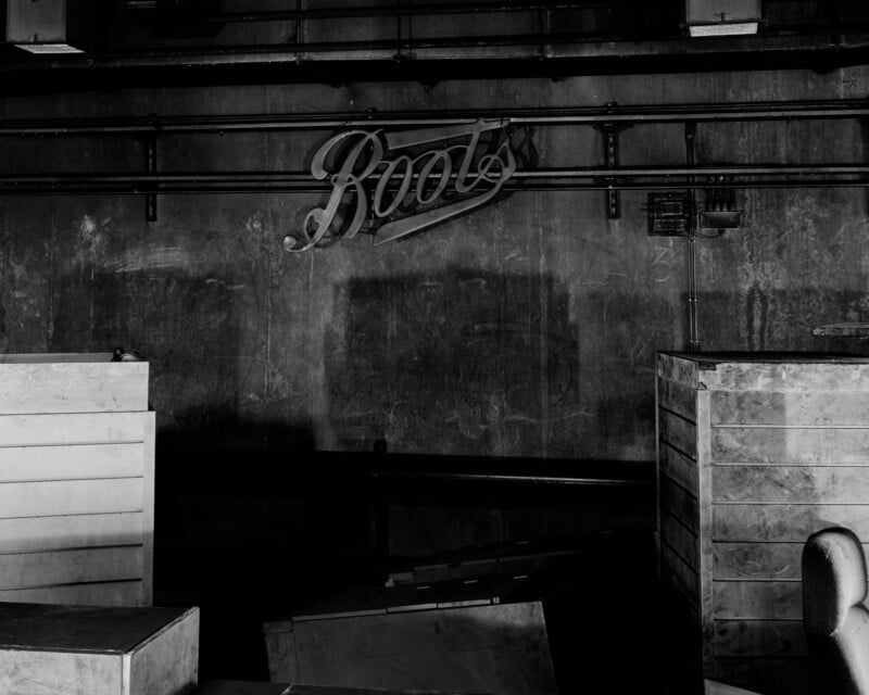 Black and white photo of an empty, dimly lit room with a vintage "Boots" sign on the wall, wooden crates, and some industrial piping.