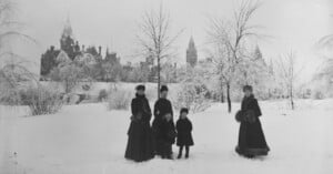 Four women and two children in dark winter clothing stand on a snowy landscape with bare trees; a large, ornate building with spires is visible in the background under a cloudy sky.