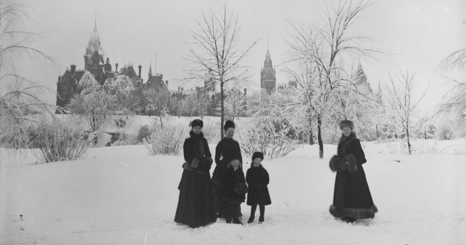 Four women and two children in dark winter clothing stand on a snowy landscape with bare trees; a large, ornate building with spires is visible in the background under a cloudy sky.