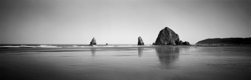 A black and white panoramic view of a sandy beach with several large sea stacks rising from the water under a clear sky.