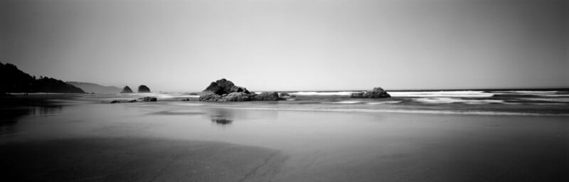 A black and white panoramic view of a sandy beach with large rock formations in the surf under a clear sky.