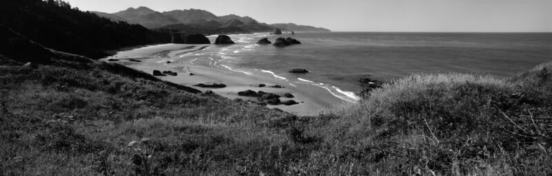 A black and white panoramic view of a sandy coastline with rocky outcrops, ocean waves, and grassy hills under a clear sky.