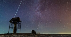 Meteors streak across a star-filled night sky above an observatory and equipment, with the Milky Way visible in the background.