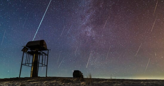 Meteors streak across a star-filled night sky above an observatory and equipment, with the Milky Way visible in the background.