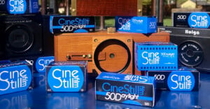 Several CineStill 50D film boxes are arranged on a blue surface with various cameras, including wooden and black plastic models, in the background. The scene is set outdoors in bright sunlight.