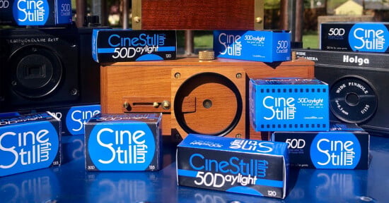 Several CineStill 50D film boxes are arranged on a blue surface with various cameras, including wooden and black plastic models, in the background. The scene is set outdoors in bright sunlight.