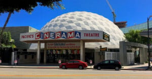 The Pacific’s Cinerama Theatre features a distinctive white geodesic dome, a retro marquee, and colorful movie posters on its façade, with palm trees and a clear blue sky in the background.