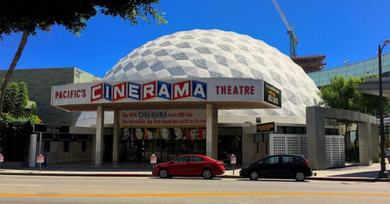 The Pacific’s Cinerama Theatre features a distinctive white geodesic dome, a retro marquee, and colorful movie posters on its façade, with palm trees and a clear blue sky in the background.