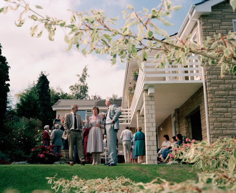 People in formal attire gather and converse in a garden outside a two-story house, with greenery and flowers present. The scene is viewed through branches in the foreground on a bright, partly cloudy day.