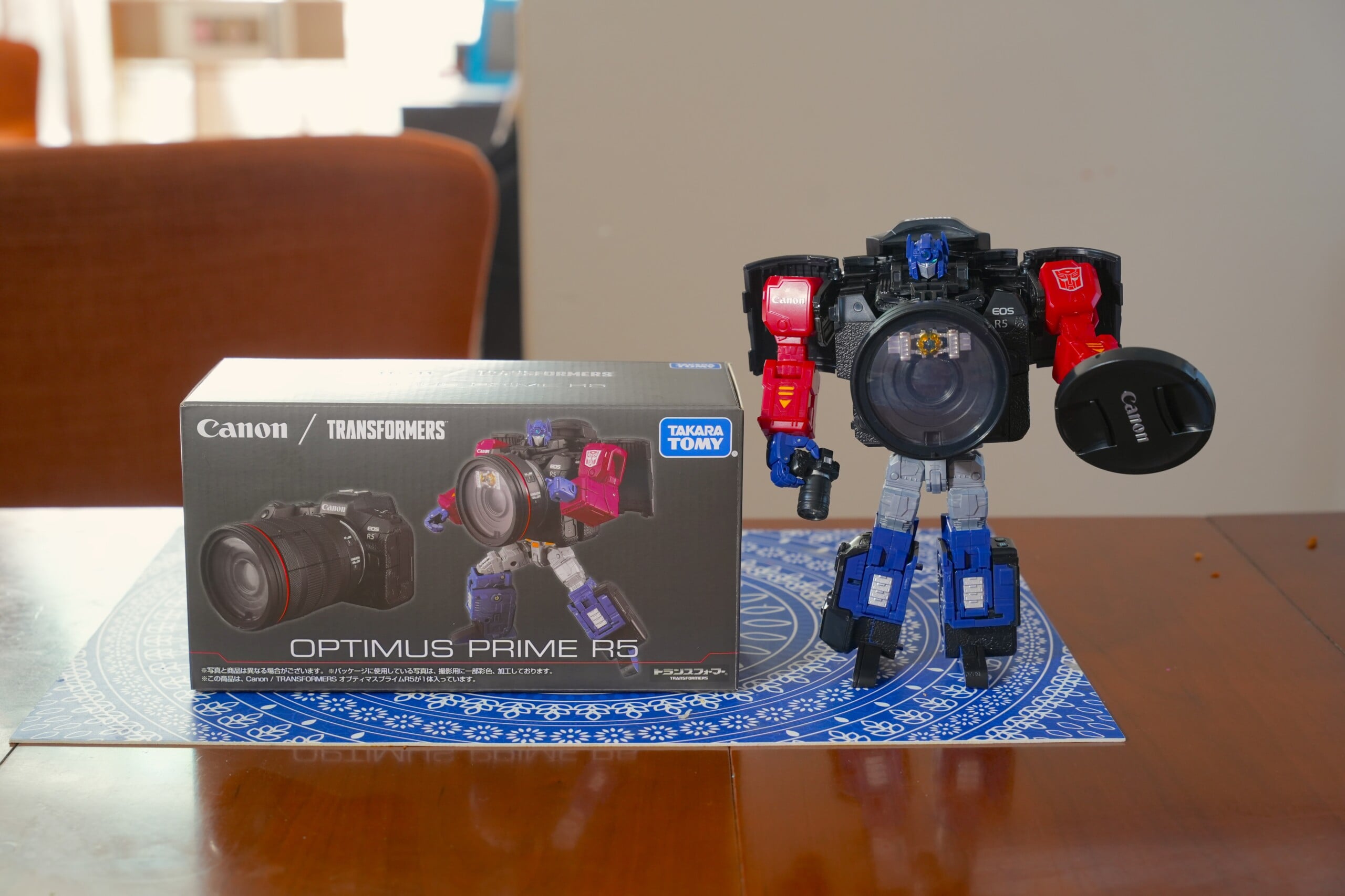 A Transformers Optimus Prime R5 action figure stands beside its Canon-themed box on a wooden table, with a camera lens as its chest and holding a lens cap in one hand.
