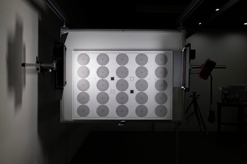 A test chart with a grid of circular star patterns is mounted on a light box in a dark room, flanked by two adjustable studio lights.