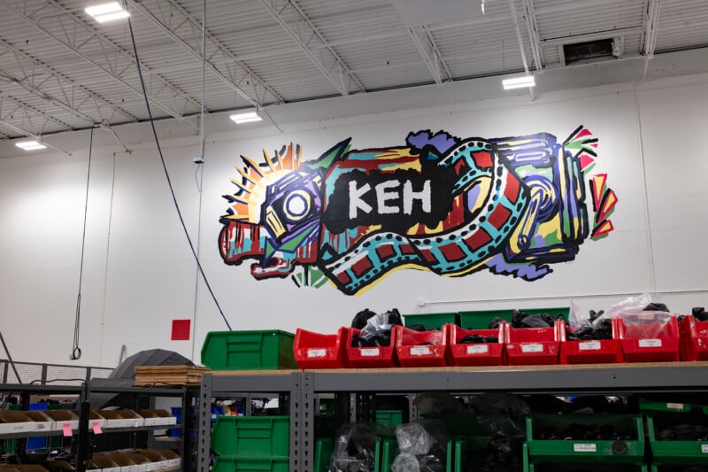 A colorful mural featuring the letters "KEH" surrounded by stylized camera and film strip imagery on a white warehouse wall above rows of storage bins.