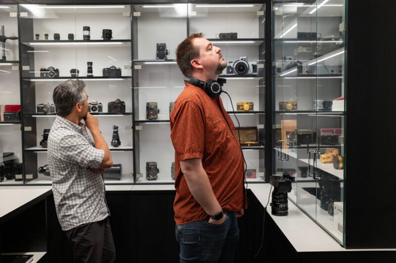 Two men stand in a shop looking at vintage cameras displayed on shelves behind glass cases.