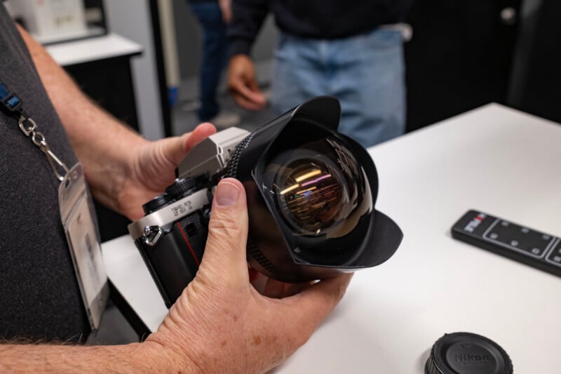 A person holds a silver and black Nikon F3T film camera equipped with a large, wide-angle lens and a petal-shaped lens hood.