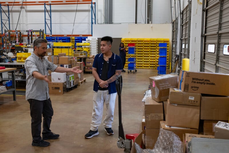 Two men stand in a warehouse talking, with stacks of cardboard boxes and shelving units filled with plastic bins in the background.