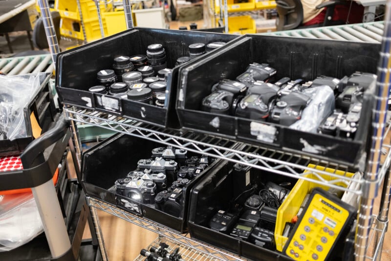 A metal shelving unit holds black plastic bins filled with various camera lenses, digital and film cameras, and electronic accessories.
