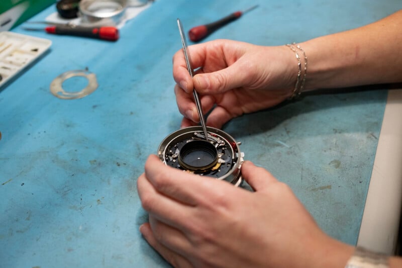 A person uses tweezers to carefully adjust the internal components of a disassembled camera lens on a blue workbench.