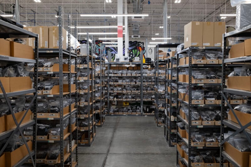 Rows of metal shelving units in a warehouse are filled with cardboard boxes and plastic-wrapped items, with a colorful mural visible on the back wall.