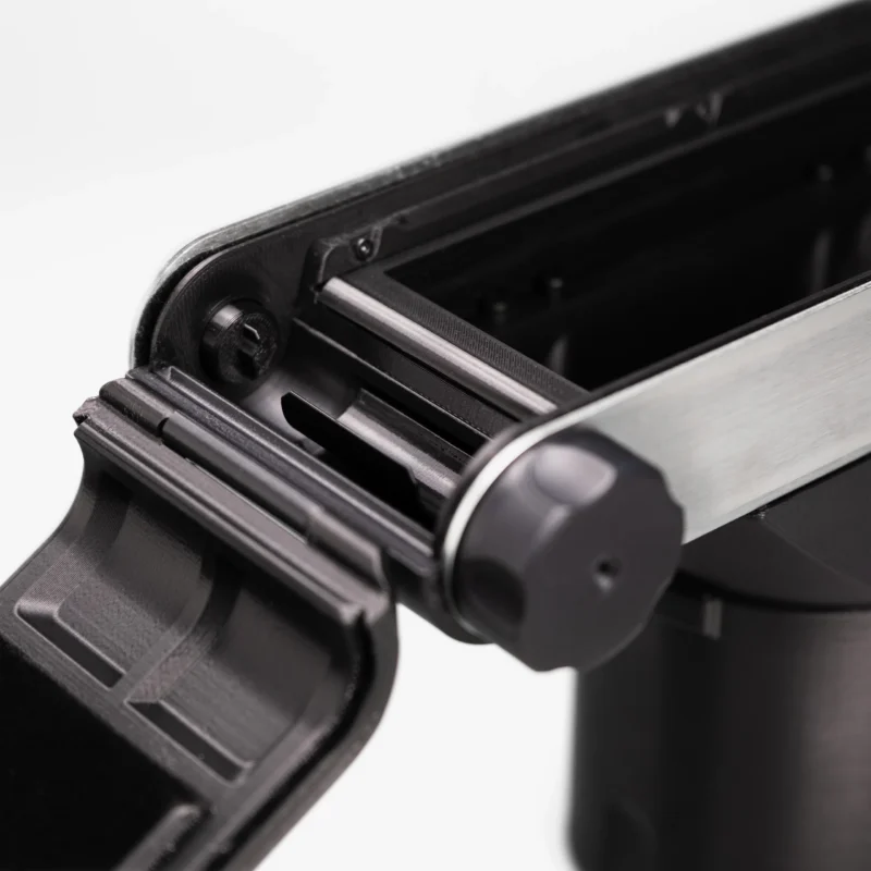 A close-up view of a black 3D-printed mechanical device with a hinged lid, a metal side rail, and a circular adjustment knob.