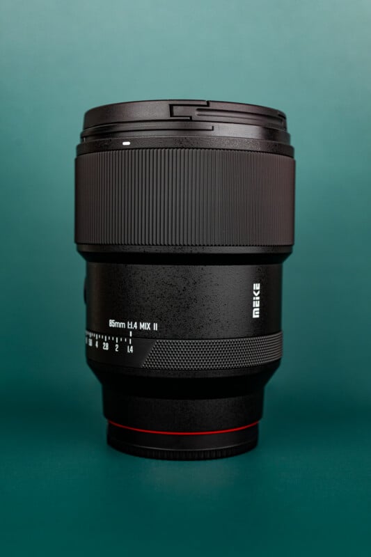 A black Meike 85mm f/1.4 camera lens stands upright on a green surface with a green background.