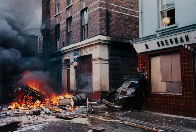 A street scene with burning debris and thick smoke. A person stands near a shopfront, taking cover. Damaged buildings and scattered wreckage line the street. The scene appears tense and chaotic.