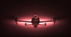 A drone with glowing red lights is captured from the front, highlighting its camera and four propellers against a dark background.
