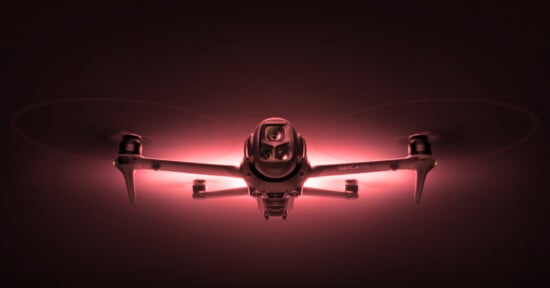 A drone with glowing red lights is captured from the front, highlighting its camera and four propellers against a dark background.