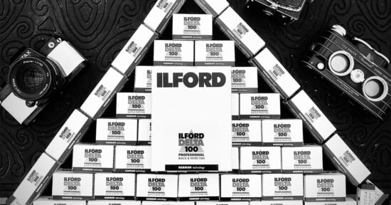 Black and white photo of stacked Ilford film boxes arranged in a triangle, with vintage film cameras placed on the patterned surface around the structure.