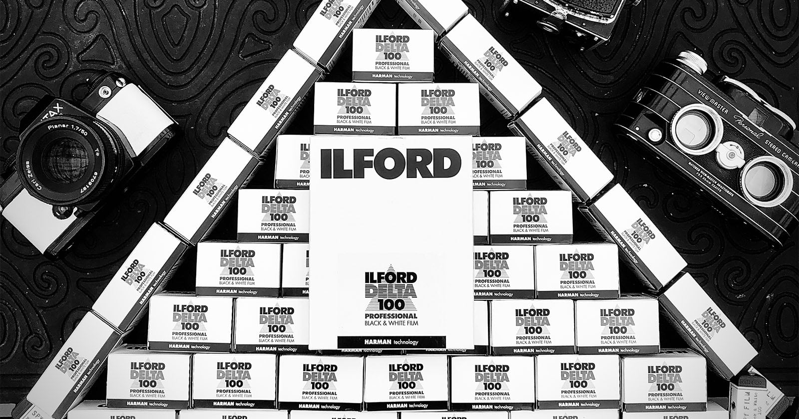 Black and white photo of stacked Ilford film boxes arranged in a triangle, with vintage film cameras placed on the patterned surface around the structure.